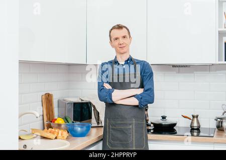 Porträt eines lächelnden, dünnen Mannes mittleren Alters mit kurzen Ingwerhaaren, blauem Langarmhemd und schwarzer Schürze, die mit gekreuzten Armen in weißem Mod steht Stockfoto