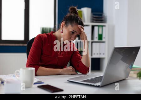 Müde Geschäftsfrau sitzt am Startup-Schreibtisch und schaut auf den Laptop-Bildschirm. Unternehmer im roten Hemd, der Kopfschmerzen im Büro hat. Inhaber von Kleinunternehmen, die Schwierigkeiten haben. Stockfoto