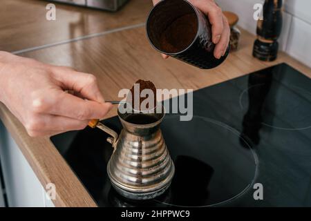 Nahaufnahme der Hände eines unkenntlichen Mannes, der einen voll gehäuften Teelöffel natürlichen gemahlenen Kaffee aus einer runden glänzenden Dose in Kupfercezve auf eine schwarze elektrische Kaffeemaschine legt Stockfoto