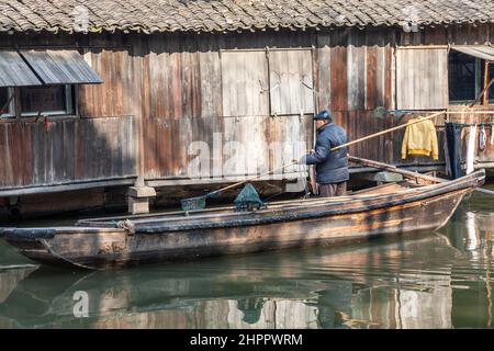 Fischer in seinem Boot, das ein Landungsnetz hält, am Fuße der Holzhäuser entlang eines Kanals im Dorf Wenzhou, China Stockfoto