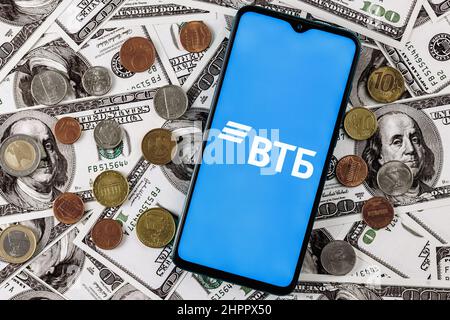 Smartphone mit VTB-Banklogo, umgeben von verschiedenen Metallmünzen auf dem Hintergrund von Dollarscheinen Stockfoto