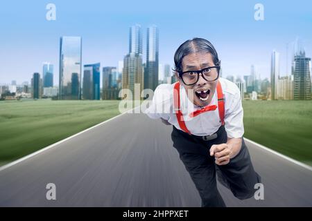Asiatischer Nerd mit hässlichem Gesicht, das auf der Straße mit Stadtansichten im Hintergrund läuft Stockfoto