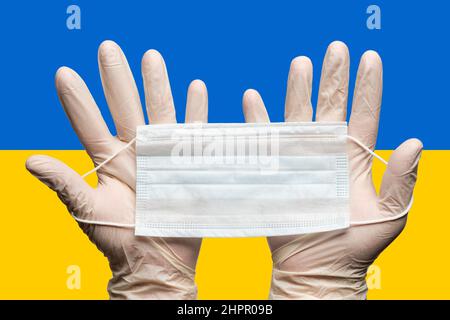 Arzt hält medizinische Gesichtsmaske in den Händen in weißen Handschuhen auf dem Hintergrund National Ukrainisch Flagge oder Flagge der Ukraine. Konzept Coronavirus-Krankheit Stockfoto