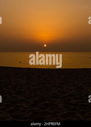 Die orange Sonne steigt langsam am Horizont während des Sonnenaufgangs auf, der Sand am Strand im Vordergrund ist noch dunkel. Stockfoto