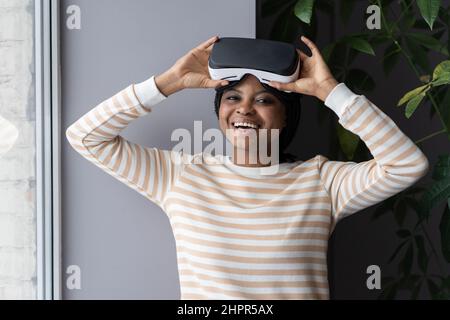 Fröhliche Afro-Frau, die den VR-Helm abnimmt, schwarze Frau, die zu Hause eine Virtual-Reality-Brille verwendet Stockfoto