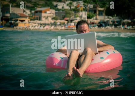 Freiberufler mit einem aufblasbaren Ring im Wasser des Meeres arbeitet am Computer. Glücklicher Mann im Schwimmkreis mit Laptop. Speicherplatz kopieren. Stockfoto