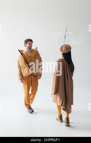 Frau in transparentem Schirm und lächelnder Mann in beiger Kugeljacke, die auf grau aufeinander zulaufen Stockfoto