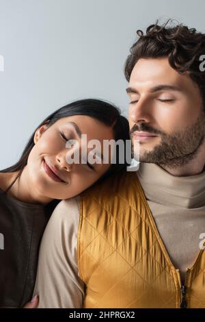 Lächelnde asiatische Frau mit geschlossenen Augen lehnt sich auf stilvolle Brünette Mann isoliert auf grau Stockfoto