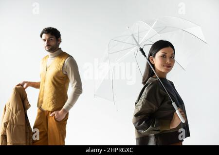 asiatische Frau lächelt unter einem transparenten Regenschirm in der Nähe eines Mannes, der eine Kugeljacke auf Grau hält Stockfoto