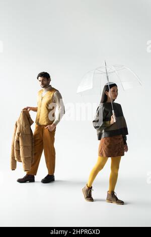 Mann mit Kugeljacke und asiatische Frau mit transparentem Regenschirm, die auf Grau läuft Stockfoto