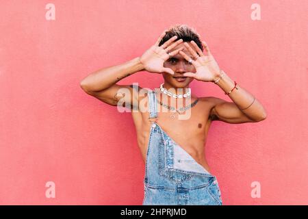 Junger, hübscher Kerl, der sein Gesicht mit Handflächen versteckt, während er die Kamera anschaut. Stilvoller Mann in Overalls, der draußen an einer rosa Wand steht. Stockfoto