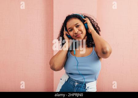 Eine Frau in Übergröße, die die Kamera mit blauen Kopfhörern an einer rosa Wand anschaut Stockfoto
