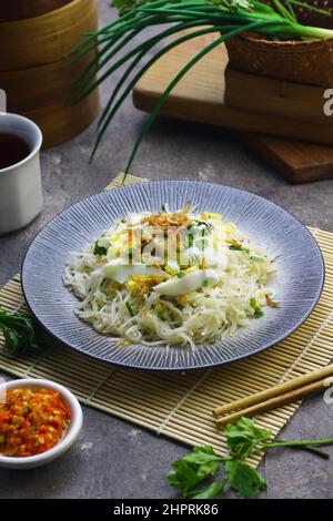 Bihun Goreng oder gebratene Reisnudeln auf Keramikplatte serviert, Belag mit Scheiben gekochtes Ei mit Sambal. Grau körnigen Hintergrund. Stockfoto