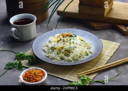 Bihun Goreng oder gebratene Reisnudeln auf Keramikplatte serviert, Belag mit Scheiben gekochtes Ei mit Sambal. Grau körnigen Hintergrund. Stockfoto