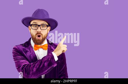 Ein Mann mit einem überraschten Gesichtsausdruck zeigt seinen Zeigefinger auf den Kopierbereich auf violettem Hintergrund Stockfoto