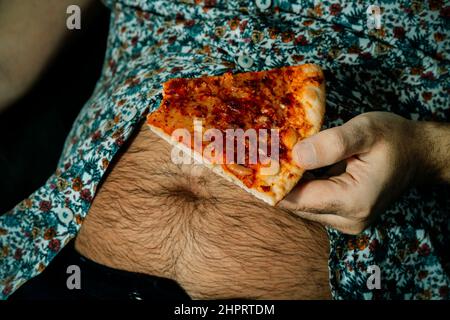 Ein kaukasischer Mann mit einem haarigen Bierbauch isst eine Portion Pizza, während er auf dem Sofa sitzt Stockfoto