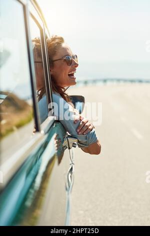 Sich auf offener Straße frei fühlen. Eine kurze Aufnahme einer attraktiven Frau, die beim Roadtrip aus einem Autofenster hängt. Stockfoto