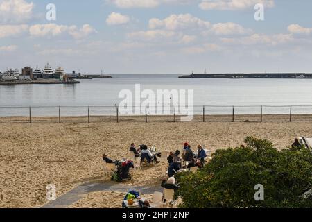 Blick auf die Küste mit Menschen, die an einer Strandbar sitzen, und dem Hafen im Hintergrund an einem sonnigen Wintertag, Sanremo, Ligurien, Italien Stockfoto