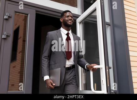 Selbstbewusster afroamerikanischer Geschäftsmann in formellem Anzug, der aus dem modernen Bürozentrum herauskommt und lächelt Stockfoto