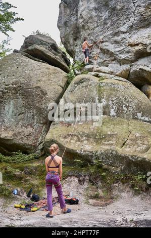 Weibliche Kletterin, die beim Klettern im Freien mit einem Seil die Führerin festsetzt. Rückansicht einer jungen Frau, die zu seinem Seilpartner schaut. Konzept von Teamwork, Vertrauen, Extremsport und Outdoor-Aktivitäten. Stockfoto