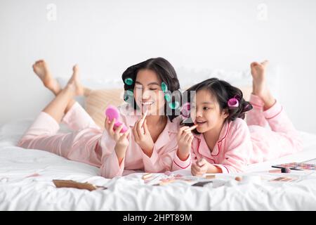 Glückliche junge japanische Frau und Teenager-Mädchen in Pyjamas und Lockenwickler liegen mit Kosmetik auf dem Bett, Spaß haben Stockfoto