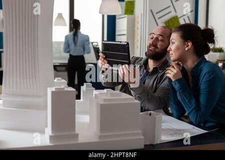 Zwei Architekten arbeiten zusammen und betrachten ein Tablet mit Blaupausen, das am Schreibtisch eines modernen Architekturbüros sitzt. Projektingenieure, die Teamarbeit mit einem digitalen Gerät neben dem Tisch mit Scale-Modell machen. Stockfoto