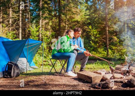 Junge Paare von Reisenden genießen Lagerfeuer im Sommerwald. Touristen sitzen am Zelt umarmt. Camping im Wald Stockfoto