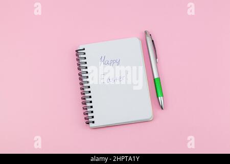 Notizblock und Stift auf rosa Hintergrund. Schriftzug Frohe Ostern. Notizbuch für Ideen, Mitteilungen, Listen und Inspirationen. Draufsicht, flach liegend mit Kopierplatz. Mock Stockfoto