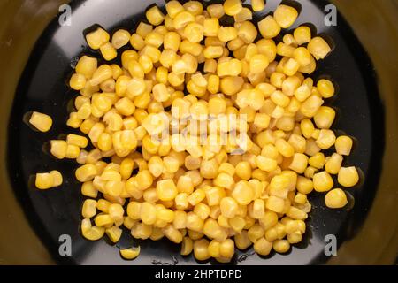 Leuchtend gelbe, süße Kerne aus Maiskonserven in einer schwarzen Keramikplatte, Makro, Draufsicht. Stockfoto