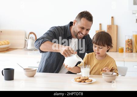 Glücklicher fürsorglicher Vater, der frische Milch ins Glas gießt, entzückender kleiner Junge, der Papa in der Küche hilft, leeren Raum Stockfoto