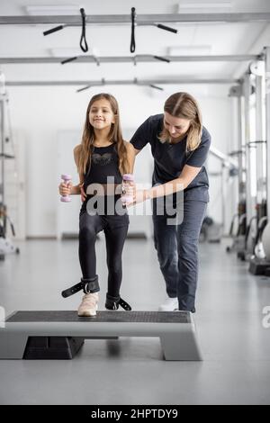 Reha-Spezialist hilft kleinen Mädchen, Übungen im Fitnessstudio zu tun Stockfoto