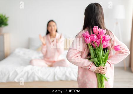 Teen Mädchen halten Strauß von Tulpen hinter dem Rücken und gratuliert asiatische tausendjährige Frau im Schlafzimmer Innenraum Stockfoto