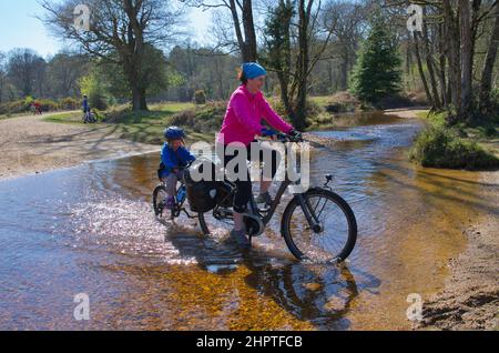 Frau auf einem Elektrofahrrad und Kind auf einem Tagalong (Anhängerfahrrad), der durch Fletchers Water ford im New Forest National Park fährt Stockfoto