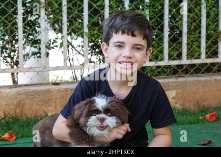 8-jähriger Brasilianer im Garten mit seinem ersten Haustier, einem Shih-Tzu-Welpe. Stockfoto