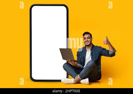 Cooler junger Araber, der mit einem Laptop in der Nähe eines riesigen Mobiltelefons mit leerem Bildschirm sitzt und den Daumen hochzeigt, ein Nachbau Stockfoto