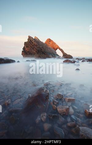 Landschaft von natürlichen Meeresbogen und Steinen im Vordergrund mit Wasser bedeckt. Berühmte Felsformation an der Moray Coast, Schottische Highlands, Schottland. Bow Fiddle Rock bei Sonnenuntergang, Langzeitbelichtung. Stockfoto