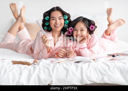 Lächelnd glücklich chinesischen weiblichen und Teenager-Mädchen in Pyjamas und Lockenwickler liegen mit Kosmetik und Maniküre Ausrüstung Stockfoto