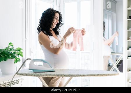 Junge hübsche Schwangerin mit lockigen Haaren bügelt die Kleidung ihres zukünftigen Babys und bereitet das Zimmer für das Neugeborene vor Stockfoto