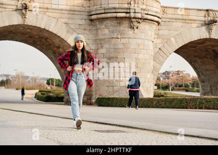 Junge Latina beim Spaziergang in der Stadt. Frau mit einem rot karierten Hemd und einer grauen Kappe, während sie mit gelben Kopfhörern Musik hört. Stockfoto