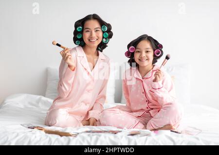 Zufriedene junge japanische weibliche und Teenager-Mädchen in Schlafanzug und Lockenwickler mit Make-up-Bürsten auf dem Bett Stockfoto