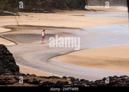 Auf der Suche nach dem Strand: Eine Frau geht allein durch einen Wellenstrom an diesem berühmten Strand, scheinbar das Wasser zu ihren Füßen suchend. Stockfoto