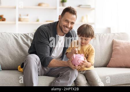 Finanzielle Kompetenz. Glücklicher kleiner Junge, der Münzen in Sparschwein legt, Vater lehrt Sohn, Einsparungen zu machen, freien Platz Stockfoto