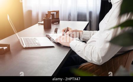 Hände, die Notizen machen, im Planer am Schreibtisch mit Laptop schreiben. Stockfoto