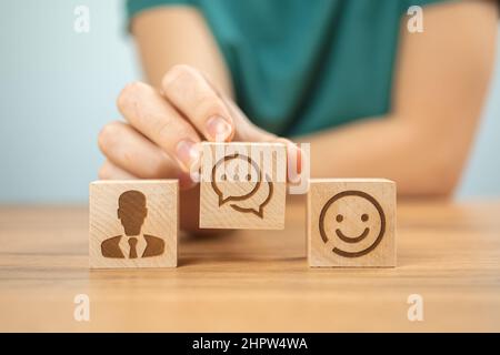 Hintergrund des Kundendienstkonzepts des Kundencenters. Hilfe und Unterstützung, Zusammenarbeit. Holzwürfel mit lächelnden Ikonen und junge Frau Hand in Büro Foto Stockfoto