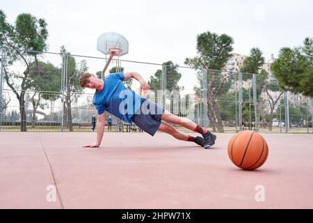 Seitenansicht eines sportlichen, jungen Mannes, der Liegestütze mit einem Basketball-Workout im Freien auf dem Basketballplatz macht. Horizontale Aufnahme. Stockfoto
