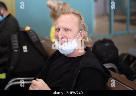 Ein blonder Europäer sitzt am Flughafen in einer bis zum Kinn heruntergelassenen Schutzmaske und lächelt. Vermeidung von Covid an überfüllten Orten. Stockfoto