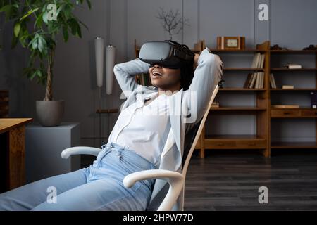 Verblüffte schwarze Geschäftsfrau, die VR am Arbeitsplatz verwendet und sich in einer entspannenden virtuellen Umgebung im Büro ausruht Stockfoto