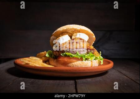 Hochwertiger Rindfleisch-Burger mit Ziegenkäse, karamellisierten Zwiebeln in einem rustikalen Brötchen mit pommes frites auf der Seite. Lecker Burger serviert auf einem rustikalen Teller auf Holztisch. Hochwertige Fotografie Stockfoto