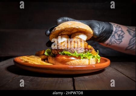 Nahaufnahme eines nicht erkennbaren Küchenchefs, der einen hochwertigen Rindfleisch-Burger mit Ziegenkäse, karamellisierten Zwiebeln, serviert mit Pommes auf einem rustikalen Teller über einem Holztisch, ausschmilbt. Hochwertige Fotografie Stockfoto