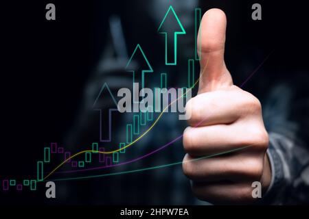 Ein Anleger mit Telefon zeigt vor dem Hintergrund einer aufsteigenden Grafik mit hoher Volatilität und gleitenden Durchschnitten einen Daumen nach oben. Bullischer Trend mit li Stockfoto